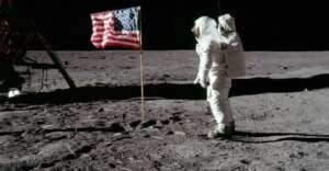 An astronaut in a white spacesuit stands on the gray, rocky surface of the Moon next to an American flag, with another part of the spacecraft visible on the left. The sky is completely black.