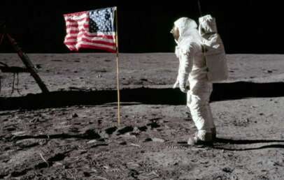 An astronaut in a white spacesuit stands on the gray, rocky surface of the Moon next to an American flag, with another part of the spacecraft visible on the left. The sky is completely black.