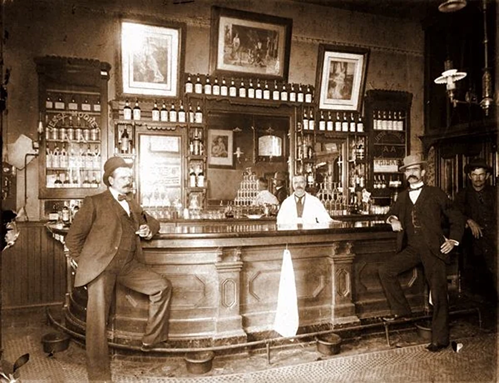 Sepia-toned photo of four men in early 1900s attire inside a bar, with ornate wooden counter, shelves of bottles, mirrors, and framed pictures behind them. Three men pose in front, one stands behind the bar.