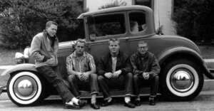 Four young men in casual 1950s attire sit and lean against a classic car with whitewall tires, parked on a street in front of a building with bushes in the background.