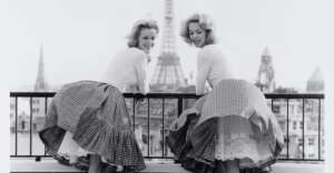 Two women in vintage dresses lean over a balcony railing, smiling toward the camera, with the Eiffel Tower and Paris cityscape visible in the background.