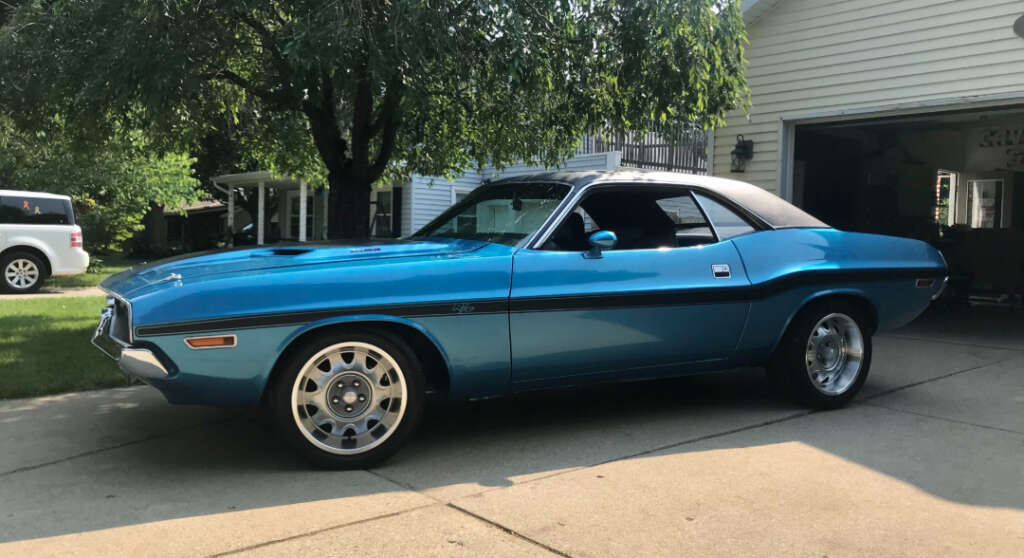 A classic blue muscle car with a black vinyl roof is parked in a driveway next to a white house and garage, with trees and another vehicle in the background.