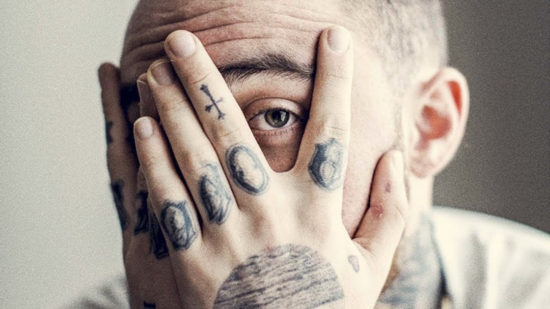 A person with tattoos on their fingers and hand covers part of their face with their hand, revealing one eye looking through their fingers. The background is blurred and neutral in color.
