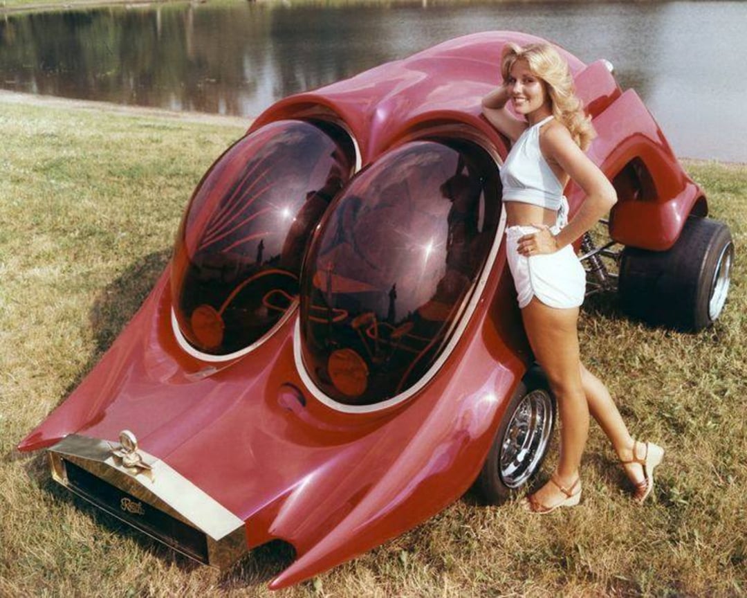 A woman in a white crop top and shorts poses beside a futuristic red custom car with large, dark bubble-shaped windows, resembling insect eyes, parked on grass near a body of water.