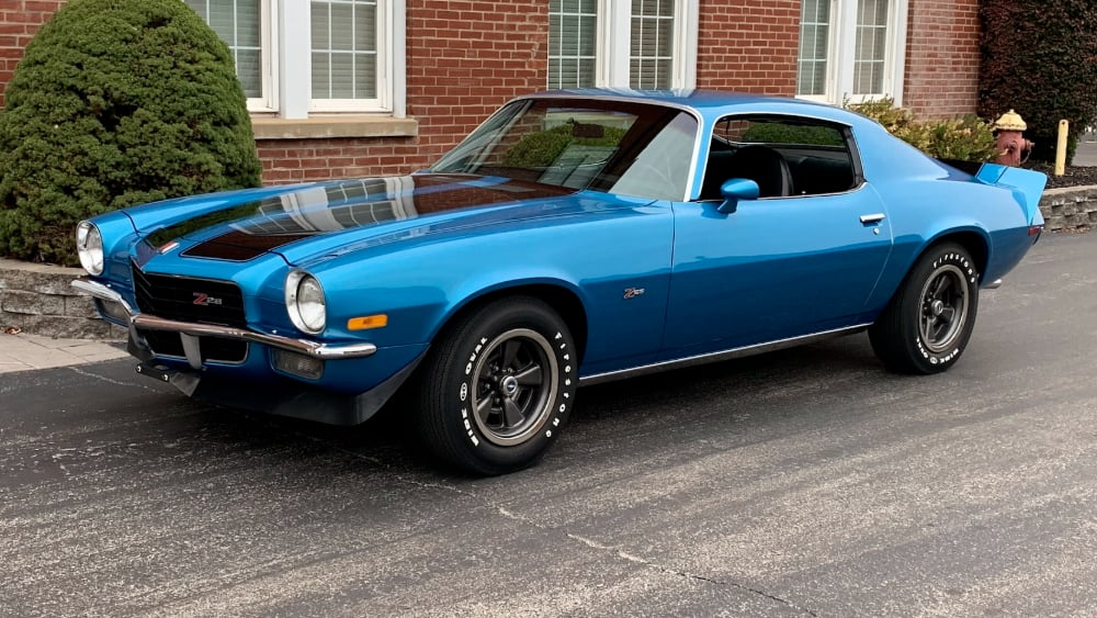 A classic blue Chevrolet Camaro Z28 with black racing stripes is parked on a street in front of a brick building and neatly trimmed bushes.