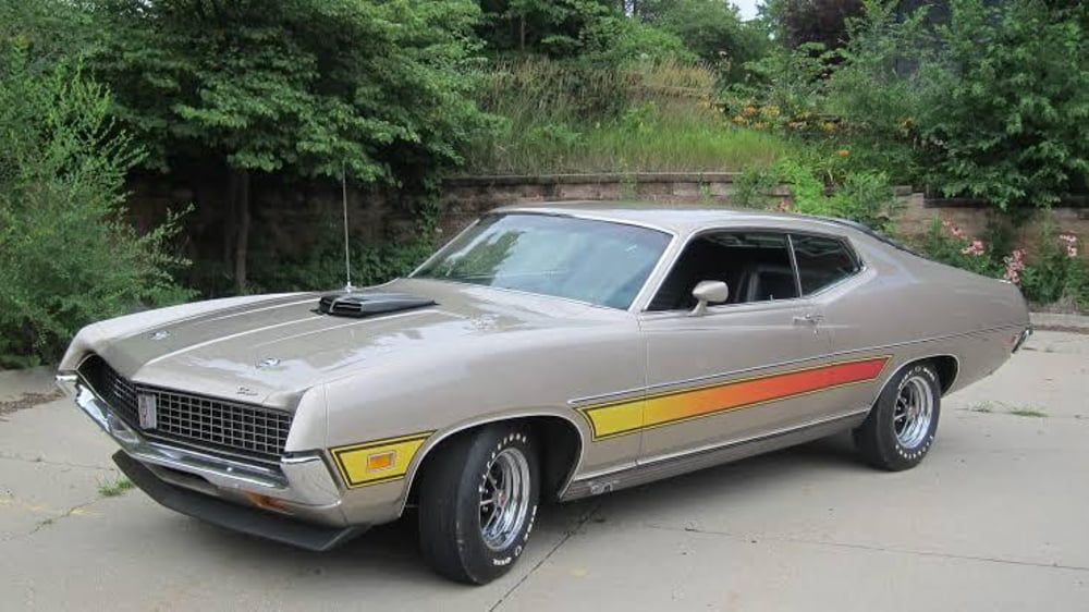 A vintage beige muscle car with a black hood scoop, chrome wheels, and yellow-to-orange gradient stripes on the sides, parked on a driveway with greenery and trees in the background.