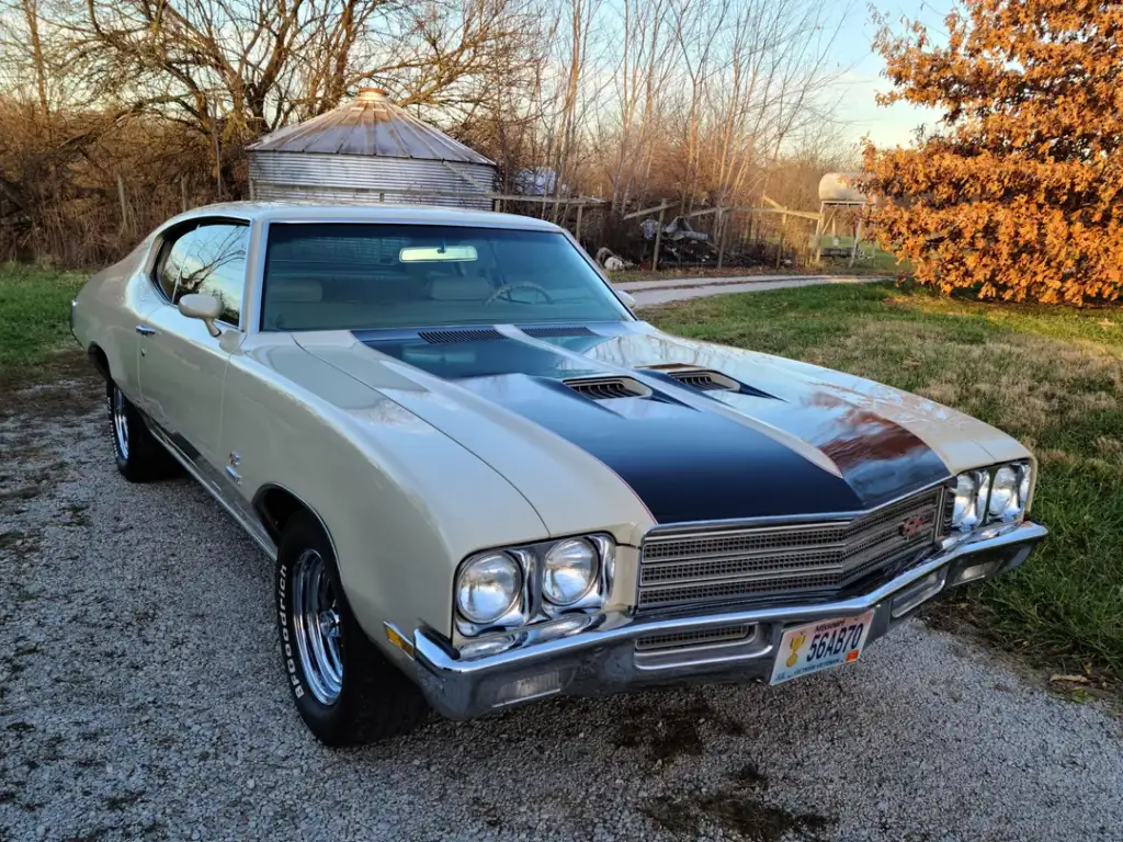 A vintage cream and black muscle car is parked on a gravel driveway near a grassy yard, with a silo and trees in the background on a sunny day.