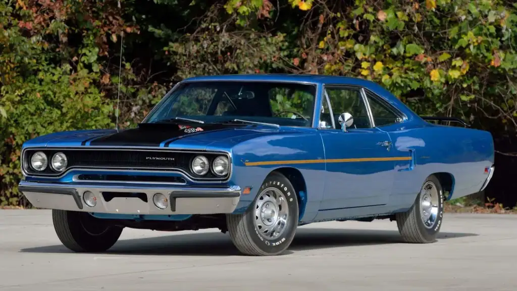 A classic blue Plymouth Road Runner muscle car with a black hood and chrome accents is parked on a concrete surface, with trees and foliage in the background.