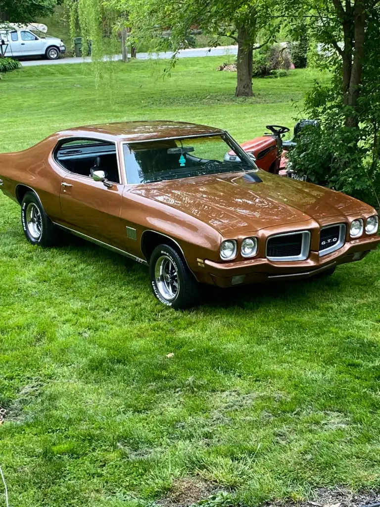 A classic brown Pontiac GTO muscle car is parked on a green lawn, surrounded by trees and bushes on a sunny day.
