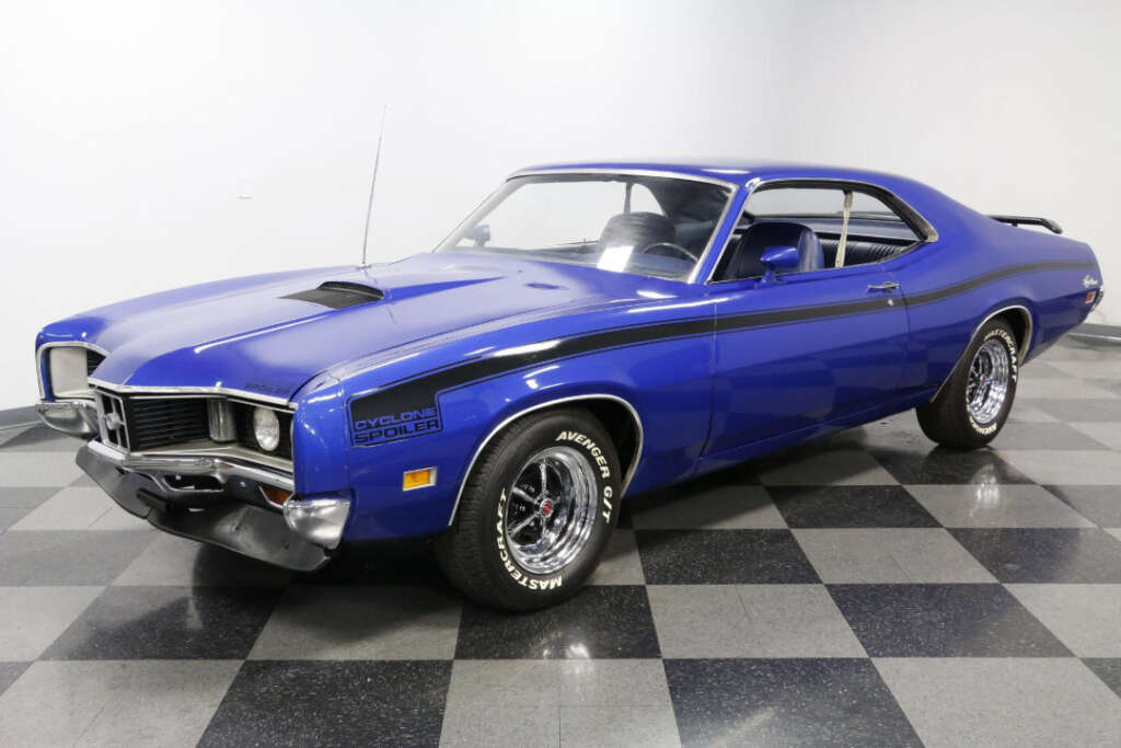 A classic blue muscle car with black detailing, chrome bumpers, and “Cyclone Spoiler” decal on the front fender is parked indoors on a checkered floor, facing left.