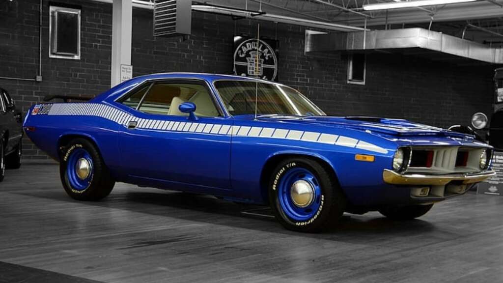 A classic blue muscle car with white racing stripes is parked indoors on a polished wooden floor, with a black brick wall and automotive decor in the background.