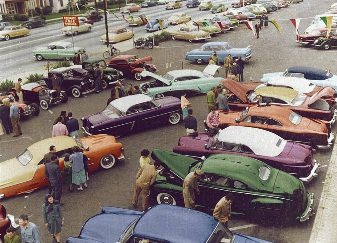 A busy 1950s car dealership lot with people walking around and examining colorful classic cars. More vintage cars and a street are visible in the background.