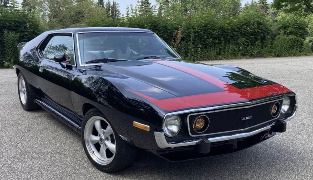 A vintage black muscle car with a red stripe down the center of the hood is parked on a paved road, surrounded by greenery and trees. The car has round headlights and silver wheels.