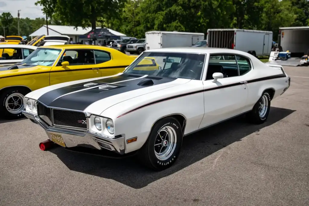 A white classic muscle car with black racing stripes is parked at a car show, with other vintage cars and trees visible in the background.