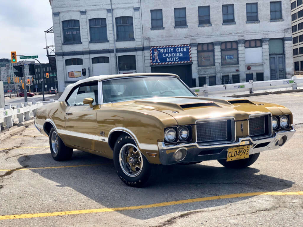 A gold vintage convertible car with a white roof is parked in an urban lot. An old brick building and a sign reading "Nutty Club Candies Nuts" are visible in the background.