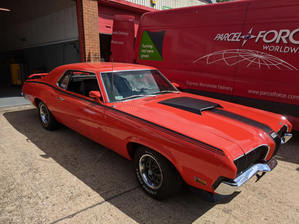 A bright red classic Mercury Cougar muscle car is parked outside near a red ParcelForce Worldwide delivery van and a brick building on a sunny day.