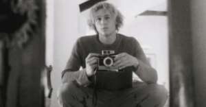 A young person with wavy hair sits cross-legged, holding a vintage camera and looking into a mirror, capturing their own reflection in black and white.