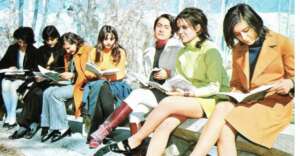 Seven young women sit on a bench outdoors, dressed in colorful 1960s or 1970s fashion, reading books together on a sunny day.