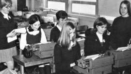 Black and white photo of five young women sitting at desks, typing on typewriters, while an older woman standing on the left observes them in a classroom setting.