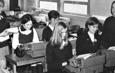 Black and white photo of five young women sitting at desks, typing on typewriters, while an older woman standing on the left observes them in a classroom setting.