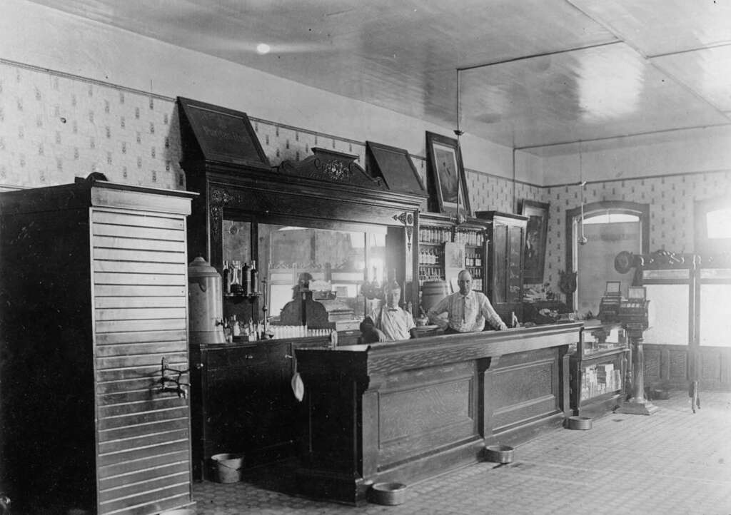 Black and white photo of an old-fashioned bar with two men standing behind a large wooden counter; shelves with bottles and glasses, patterned wallpaper, and vintage decor are visible in the background.