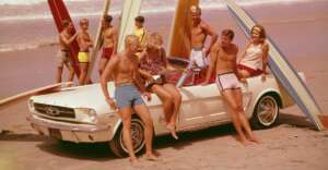 A group of young people in swimsuits gather around a white convertible on a sandy beach, holding surfboards. One person sits on the car playing guitar, while others stand nearby, with the ocean waves in the background.
