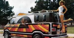 A woman in a white outfit poses on the rear ladder of a customized van with vibrant red, yellow, and orange stripes and a mural of a wizard on the side, parked outdoors near trees.