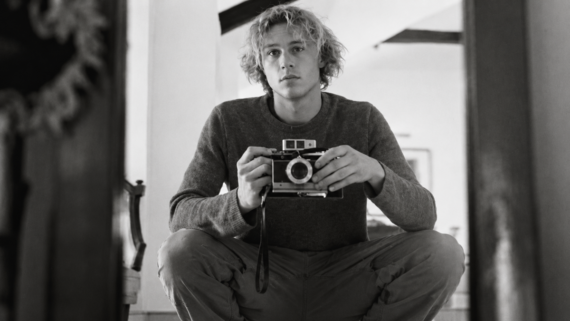 A young person with tousled hair crouches and holds a vintage camera, looking into a mirror. The black and white photo has a calm, introspective mood.