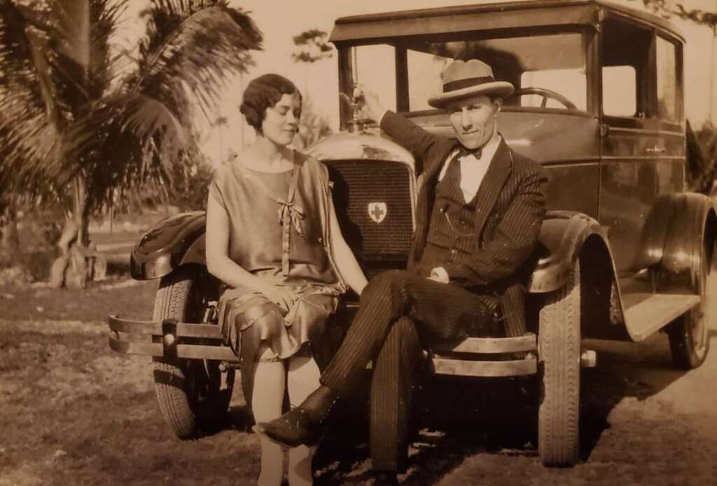A woman in a dress and a man in a suit and hat sit on the bumper of a vintage car, possibly from the 1920s, outdoors with palm trees in the background. The photo is sepia-toned and nostalgic.