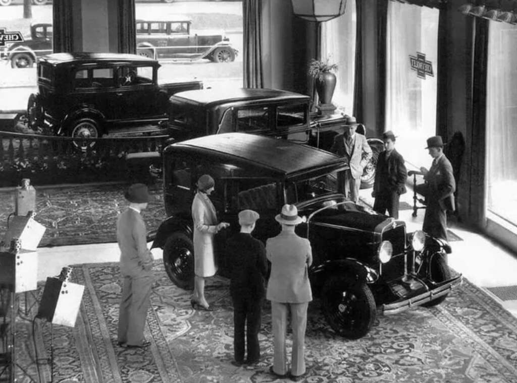 A group of people in 1920s attire gathers inside an elegant car showroom, examining vintage automobiles displayed on ornate rugs, with large windows revealing more cars outside.