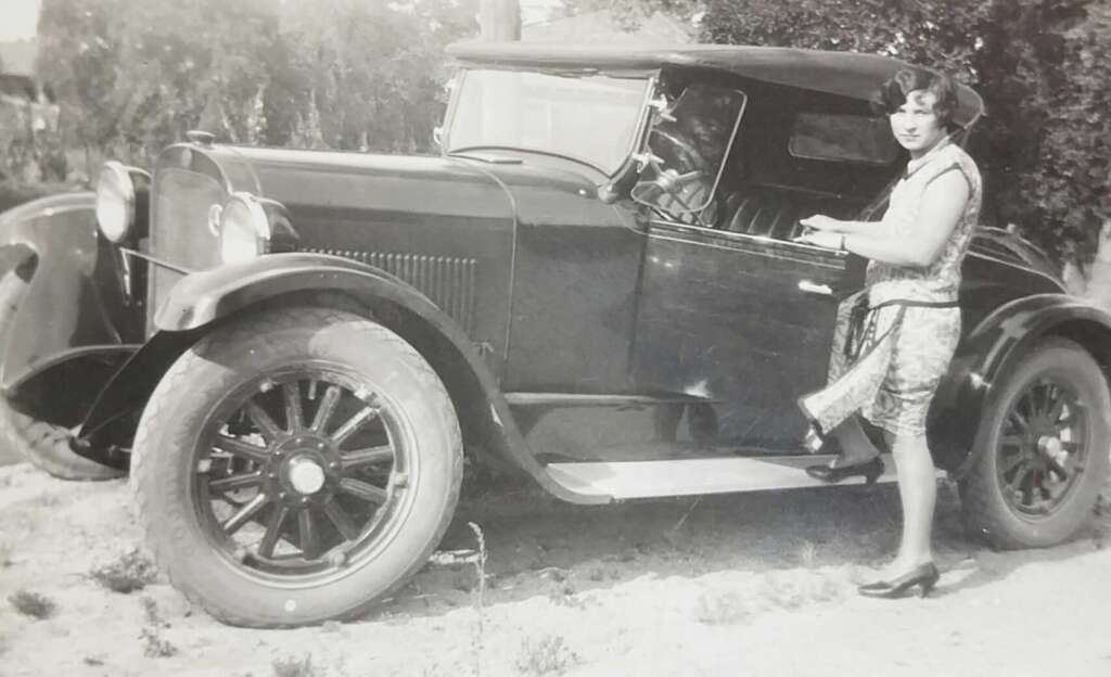 A woman in vintage clothing stands beside an old-fashioned car, holding the door handle and smiling at the camera. The image is in black and white, suggesting it is from the early 20th century.
