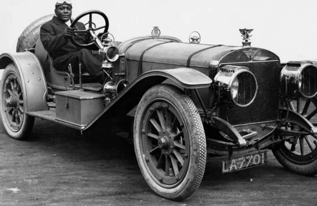 A Black man wearing driving goggles and a cap sits in the driver’s seat of a vintage open-top car with large spoked wheels and a visible front license plate reading “LA 7201.”