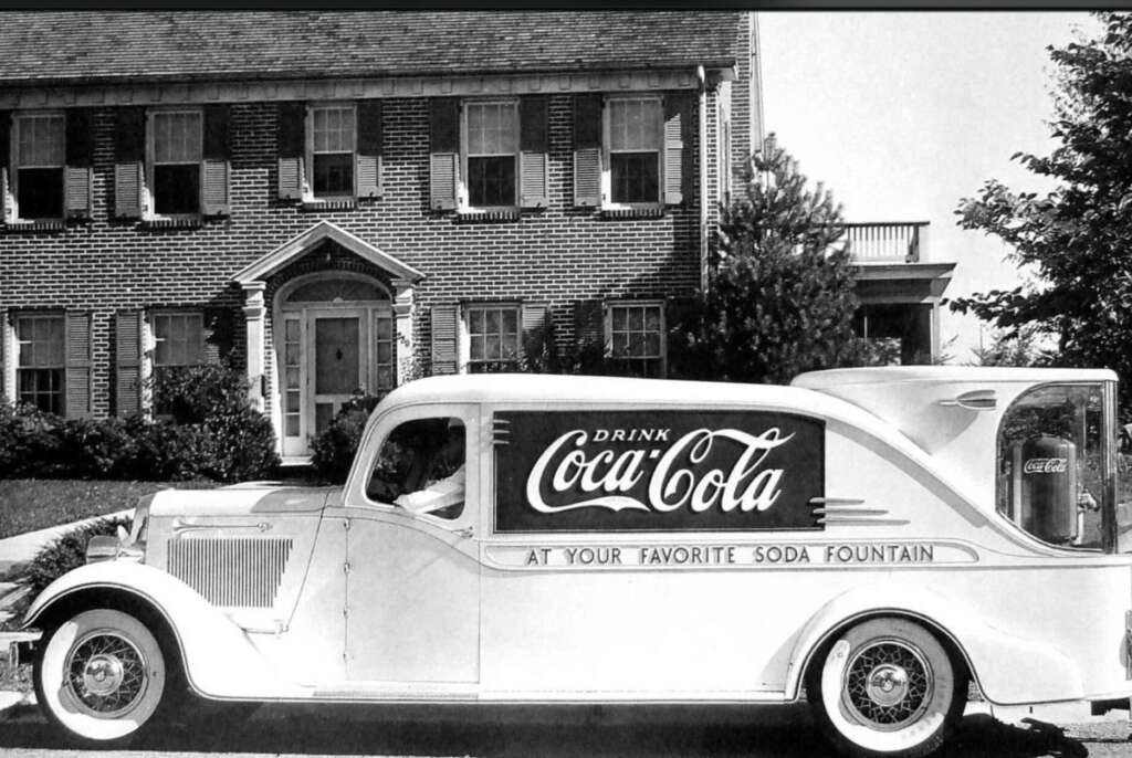 A vintage Coca-Cola delivery truck is parked in front of a two-story brick house. The truck is white with large Coca-Cola branding and the slogan "At Your Favorite Soda Fountain" on its side.
