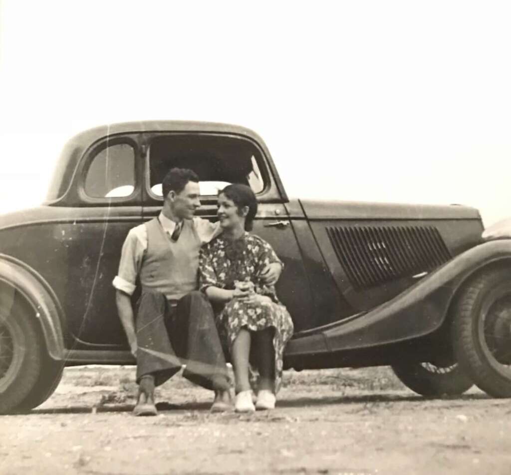 A vintage black-and-white photo of a smiling couple sitting on the ground, close together, in front of an old-fashioned car. The woman wears a floral dress, and the man is in a shirt with rolled sleeves and a vest.