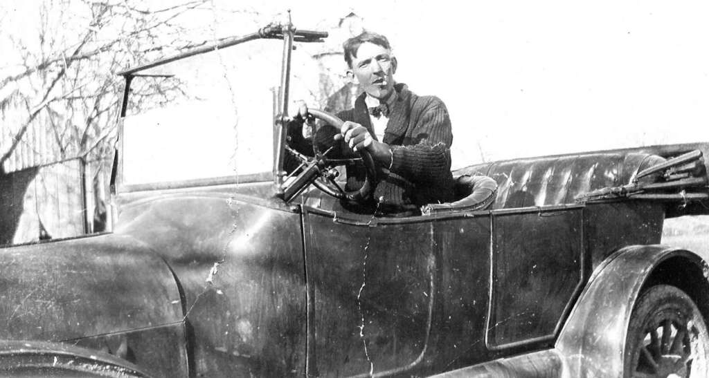 A man in vintage clothing sits behind the wheel of an old-fashioned, open-top car, looking ahead. The background shows a leafless tree and a rustic building, suggesting a rural setting. The image is black and white.