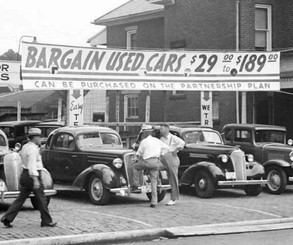 Black and white photo of a used car lot with vintage cars. A large banner advertises "Bargain Used Cars $29.00 to $189.00" and mentions a partnership plan. Several people are viewing the cars.