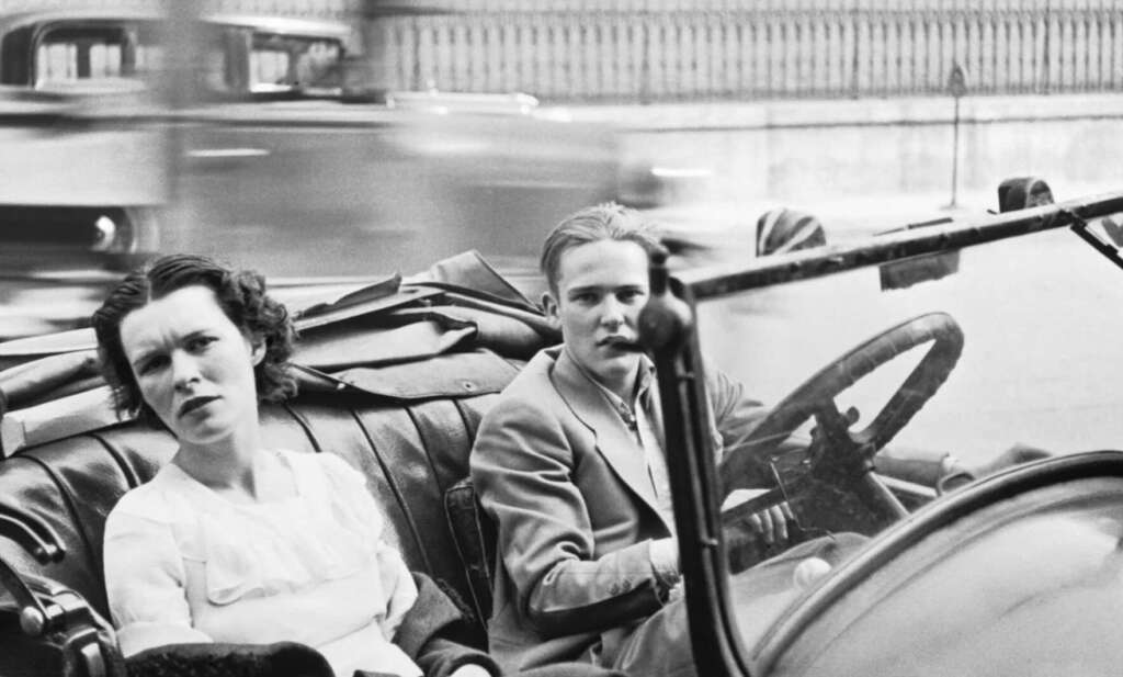 Two people sit in the front seat of a vintage convertible car, with one driving and the other looking toward the camera. Blurred cars and a fence are visible in the background, suggesting motion and a city setting.