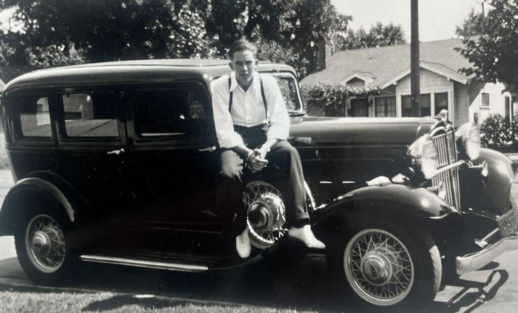 A young man in a white shirt and suspenders sits on the fender of a vintage car parked on a lawn, with a house and trees in the background. The photo is black and white.