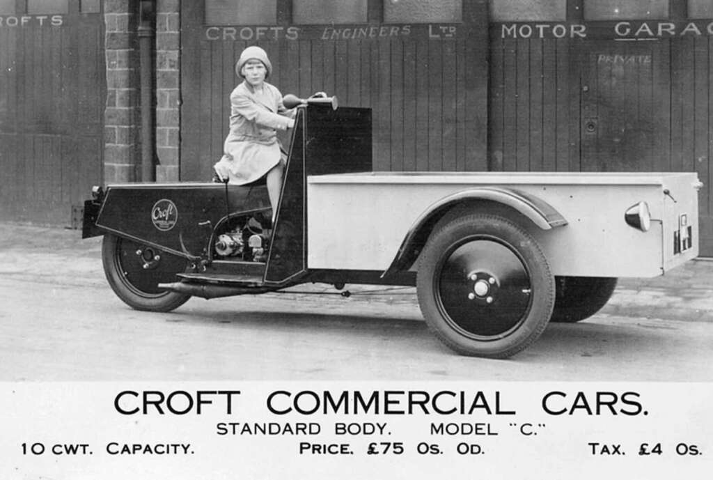 A woman sits in a vintage Croft Commercial Car with a flatbed body, parked in front of a garage. The sign advertises its model C, 10 cwt capacity, price of £75, and tax of £4 in early 20th-century style.