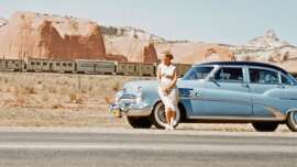 A woman in a white dress stands by a vintage blue car parked on the roadside, with rocky red desert cliffs and a train in the background under a clear sky.