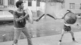 Two children play-fight with toy swords and shields on a city sidewalk. One child wears a t-shirt and jeans; the other wears a cap, t-shirt, and shorts. Parked cars and apartment buildings are in the background.