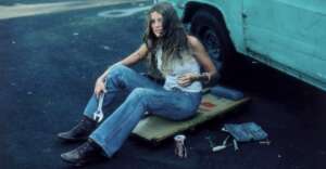 A woman in a white tank top and jeans sits on the ground by a teal van, holding a wrench. Tools, bolts, and a can of Coca-Cola are scattered nearby. She appears relaxed, with long hair and boots.