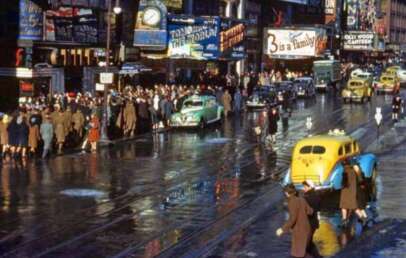A busy city street scene from the mid-20th century shows crowds of people in coats and hats, vintage cars, wet pavement, and colorful billboards, including a large sign reading “5 is a Family.”