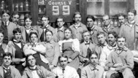 A group of men and one woman in work coats pose together in front of shelves filled with bottles and jars. A sign above their heads reads "Course V 99." The photo appears historical and black-and-white.