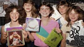 Five smiling teenage girls with 1980s hairstyles hold up Muppet-themed folders, albums, and shopping bags, standing together indoors in front of a grid wall display.