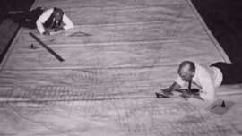 Two men in shirts and ties lie on a large floor plan, using rulers and compasses to draw and measure details on the oversized architectural blueprint spread across the floor.