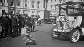 A shirtless man pulls a large vehicle with ropes held in his mouth while a crowd of onlookers watches on a city street lined with early 20th-century cars and buildings.