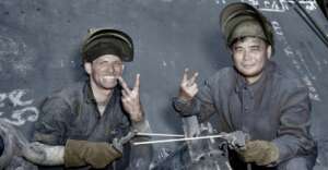 Two male workers in welding gear smile and show peace signs with their gloved hands. Both are wearing protective helmets flipped up, and their faces are marked with soot. Industrial equipment and markings are visible in the background.