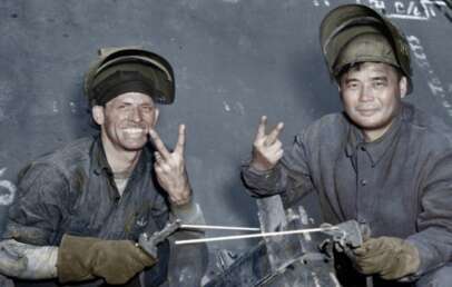 Two male workers in welding gear smile and show peace signs with their gloved hands. Both are wearing protective helmets flipped up, and their faces are marked with soot. Industrial equipment and markings are visible in the background.