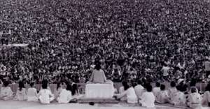 A black-and-white photo shows a person seated on a stage, facing and addressing a massive crowd of people. Several others sit on the stage beside the main figure, all viewed from behind.